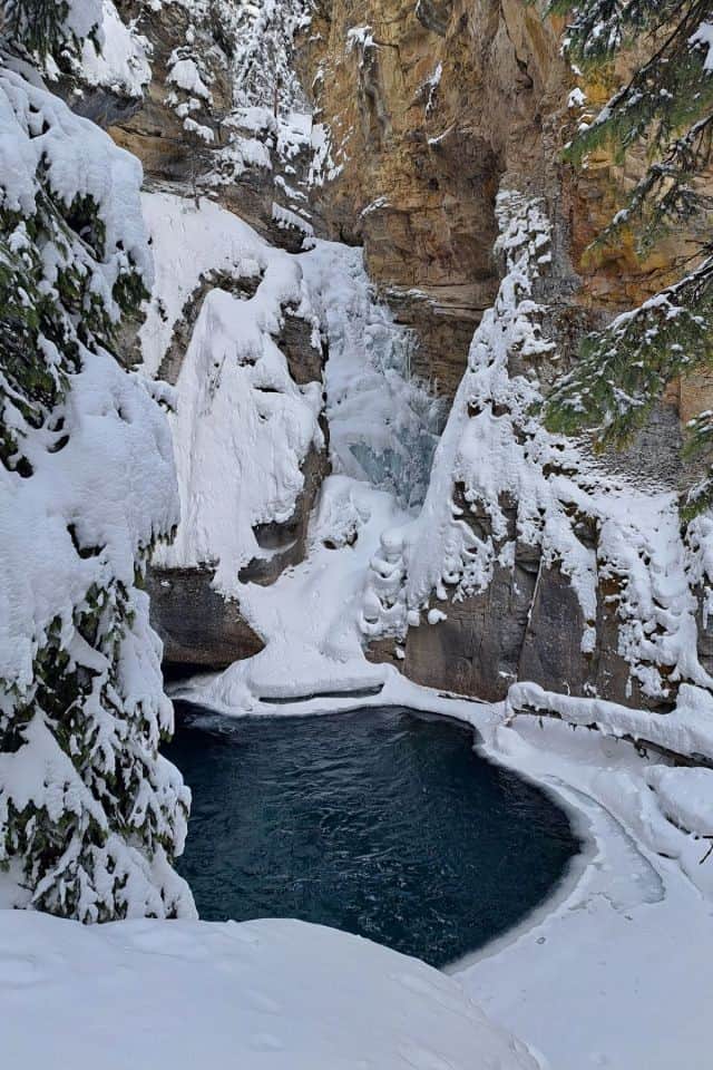 A frozen lower waterfall plunges over the cliff into a pool of blue still open water in Johnston Canyon Banff National Park Canada