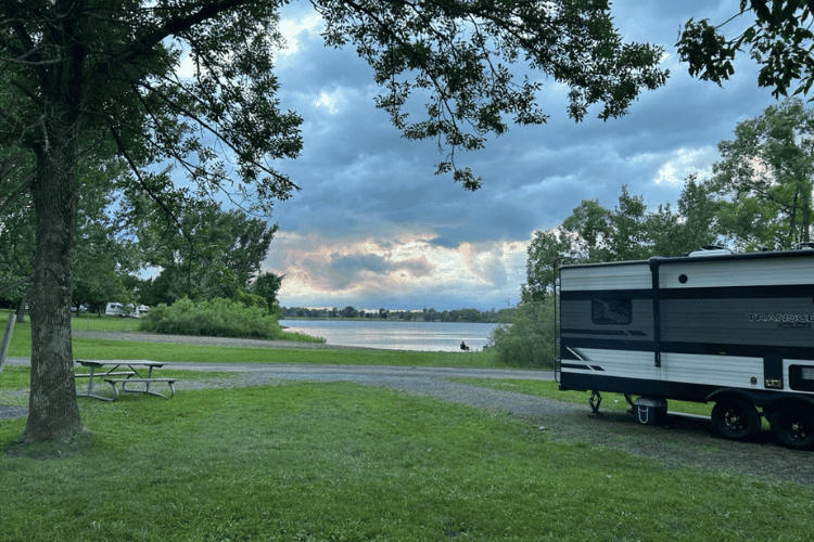 River view wth a camper trailer and picnic table in the foreground, cloudy sky.